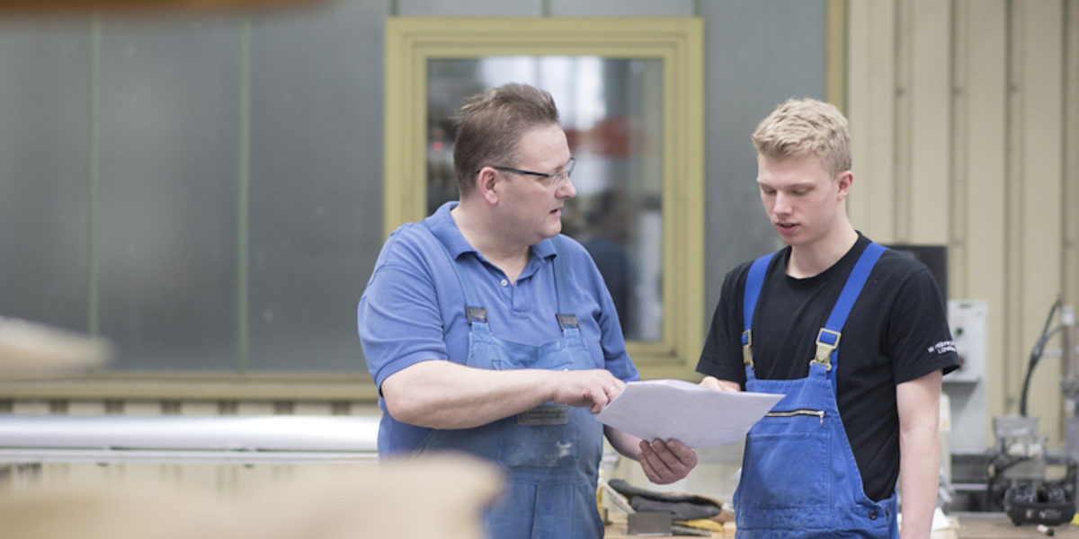 Zwei Männer in Arbeitskleidung stehen in einer Produktionshalle. Der Ältere zeigt dem Jüngeren ein Blatt Papier und spricht.