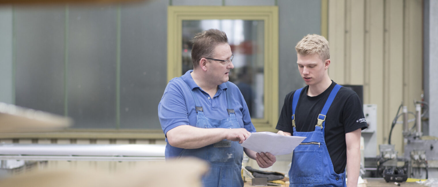 Zwei Männer in Arbeitskleidung stehen in einer Produktionshalle. Der Ältere zeigt dem Jüngeren ein Blatt Papier und spricht.