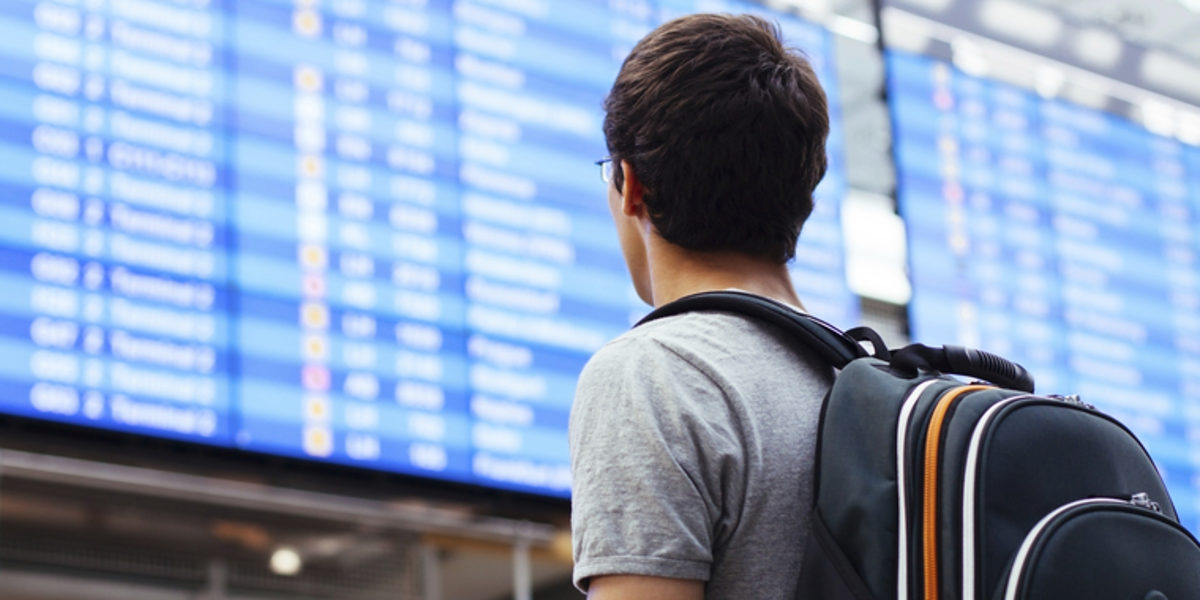 Eine Person mit einem Rucksack ist von hinten zu sehen, im Hintergrund eine Anzeigentafel wie an Bahnhöfen oder Flughäfen.