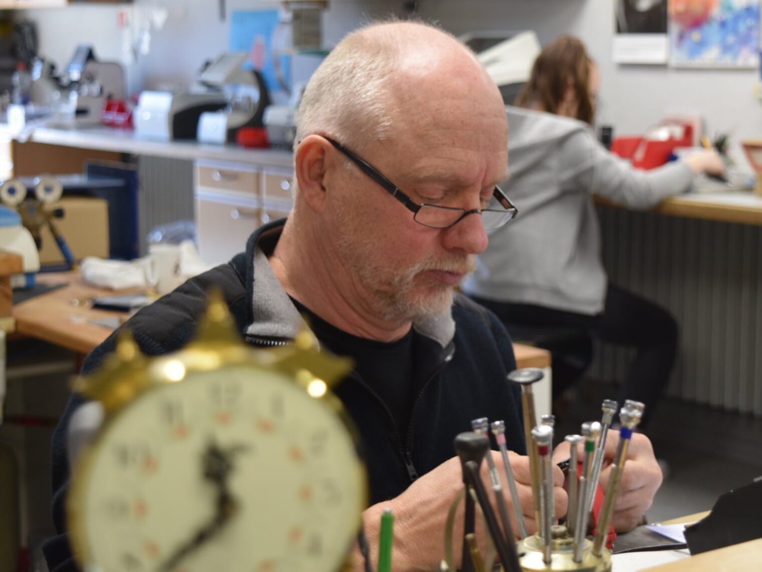 Älterer Mann mit Brille sitzt in einer Werkstatt zwischen Werkzeugen und blickt prüfend auf eine Armbanduhr in seinen Händen.