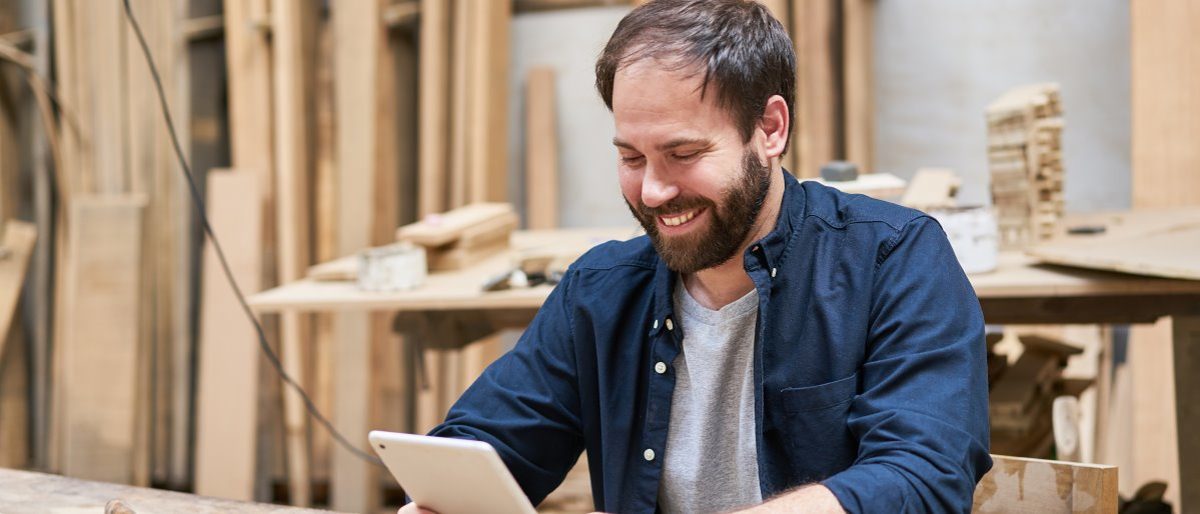 Für Digitalisierungsvorhaben können Handwerker Fördermittel beantragen. Mann sitzt in einer Schreinerwerkstatt und schaut lächelnd auf ein Tablet.