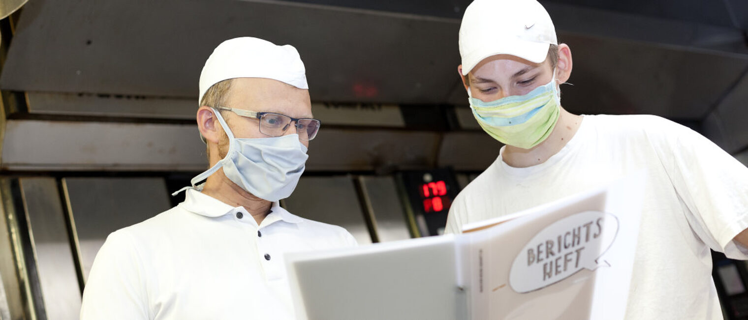 Zwei Männer in weißer Arbeitskleidung der Bäcker und Mund-Nasen-Schutz sehen in einen Ordner mit Aufschrift "Berichtsheft".