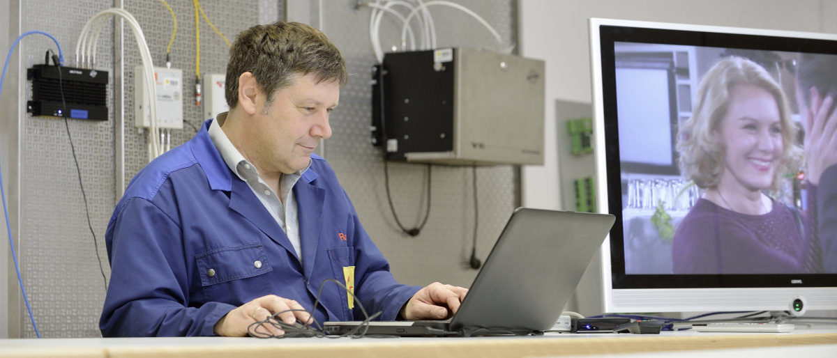 Strukturumfrage im Handwerk: Hier online teilnehmen. Mann in blauem Arbeitsjacke sitzt an Tisch vor einem und arbeitet an Laptop, Elektronik und Fernseher im Hintergrund.