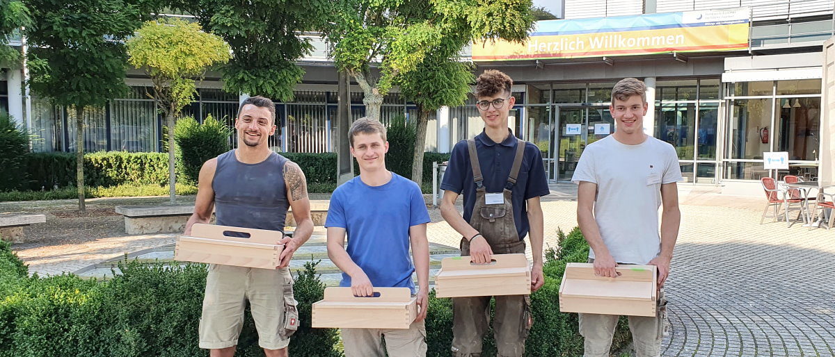 Kammersieger Felix Wilhelm (r.) mit seinen Tischler-Kollegen. Vier junge Männer stehen vor einem Gebäude im Freien. jeder hat einen Kasten aus Holz mit Griff oben in der Hand.