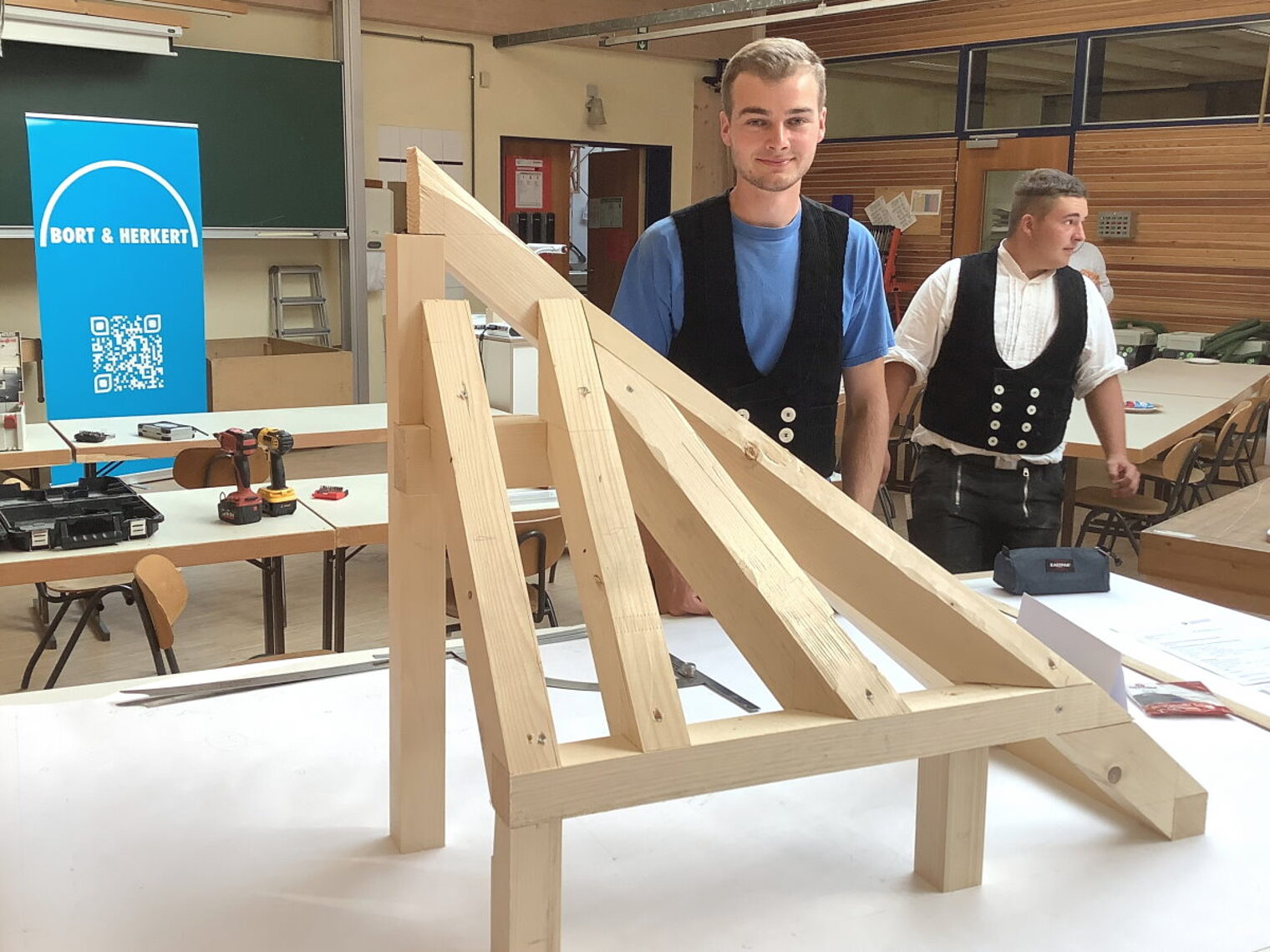Eine Holzkonstruktion steht auf einem Tisch in einem Schulraum. Dahinter steht ein junger blonder Mann in blauem T-Shirt