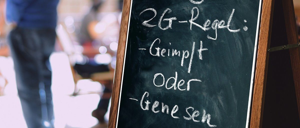 Neue Corona-Verordnung erlaubt 2G-Optionsmodell für Betriebe. Auf einer schwarzen Tafel steht mit weißer Kreide "2G-Regel: Geimpft oder Genesen. Im Hintergrund unscharf Personen zu sehen.