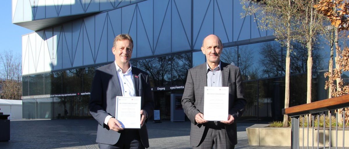 Ralf Schnörr und experimenta-Geschäftsführer Dr. Wolfgang Hansch (r.) Zwei Männer in Anzügen stehen vor einem Gebäude mit Glasfassade und halten Urkunden in DIN-A4-Format vor sich in den Händen.