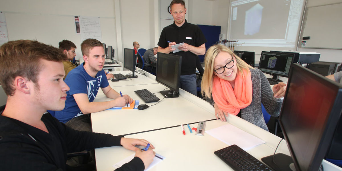 Schüler & Lehrer beim Unterricht in einem Raum mit Computern, im Vordergrund deutet eine Schülerin auf einen Monitor