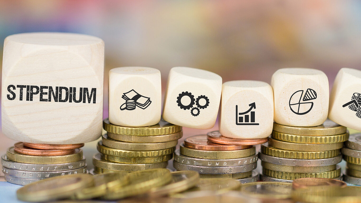 Mit einem Stipendium können Handwerker die Weiterbildung finanzieren. Auf mehreren Münzstapeln liegen Holzwürfel. Die Würfel sind mit verschiedenen Motiven zum Thema Stipendium bedruckt.