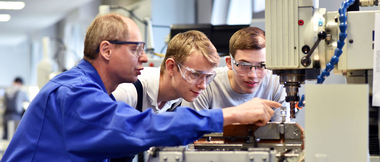 Bei Fragen helfen die Ausbildungsberater Betrieben und Azubis. Ein älterer Mann zeigt und erklärt zwei jüngeren Männern eine Maschine in einer Werkstatt.